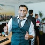 Smiling businessman showing thumbs up in a lively office environment with colleagues in the background.